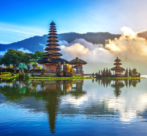 Traditioneller balinesischer Tempel am Seeufer inmitten tropischer Natur, mit Spiegelung im Wasser, umgeben von Nebelschwaden und gr&uuml;nen Bergen bei Sonnenaufgang.