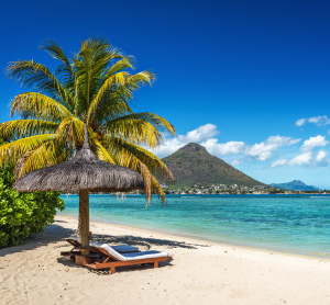 Idyllischer Sandstrand auf Mauritius mit Liege unter einer Palmen-Schilf-Sonnenschirm, t&uuml;rkisblauem Meer und Blick auf einen markanten gr&uuml;nen Berg unter strahlend blauem Himmel.