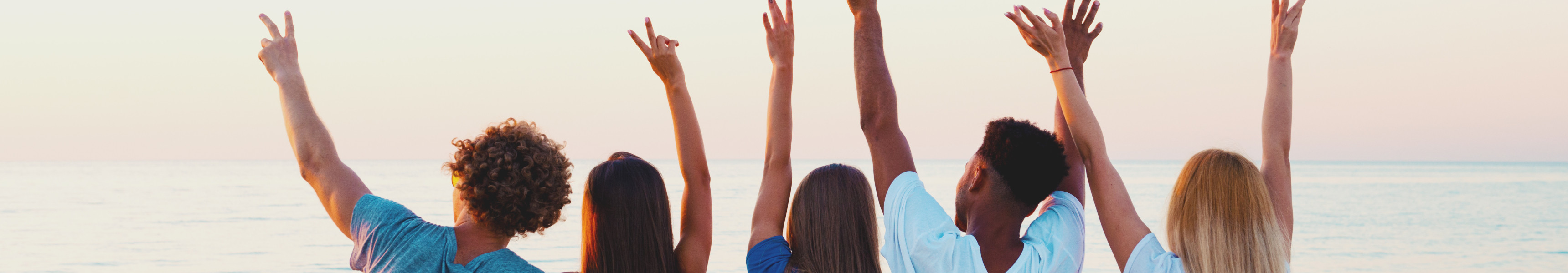 F&uuml;nf junge Erwachsene sitzen nebeneinander im Sand, schauen auf das Meer und heben freudig die Arme in die Luft.