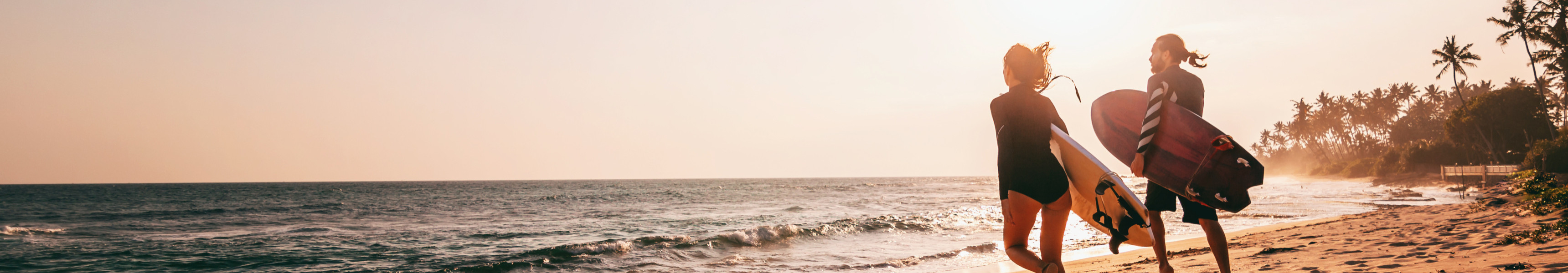 Zwei Surfer laufen mit ihren Brettern am Sandstrand entlang ins Meer, während die Sonne über dem Ozean untergeht und Palmen die tropische Kulisse ergänzen.