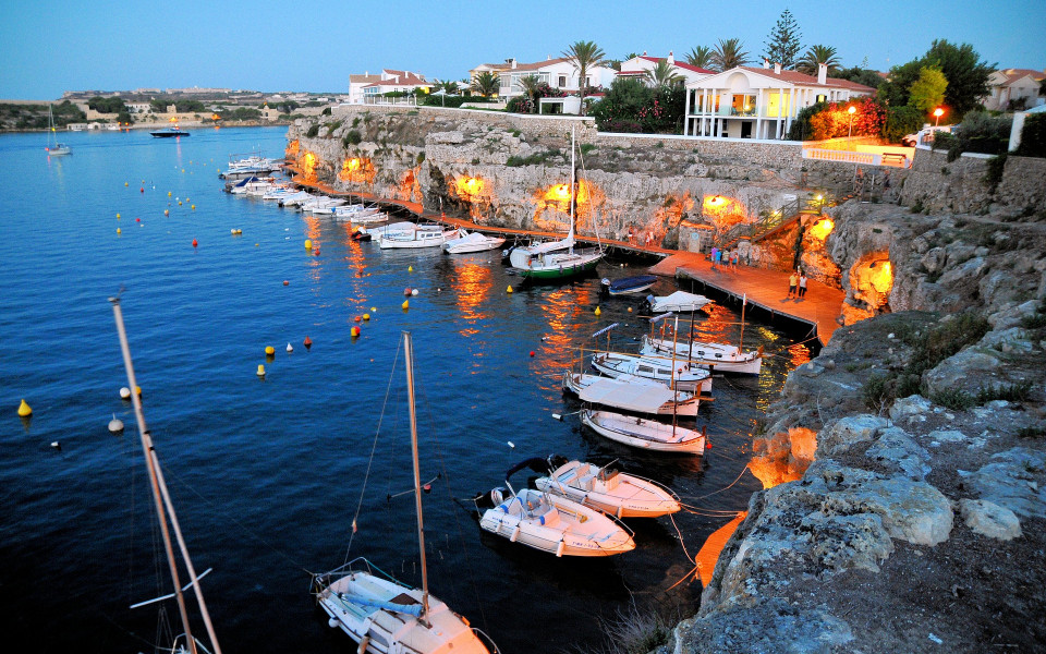 Hafen in Menorca mit Boote am Abend, umgeben von beleuchteten Felsen und wei&szlig;en H&auml;usern. Der Himmel ist leicht bew&ouml;lkt und die Atmosph&auml;re ist ruhig.