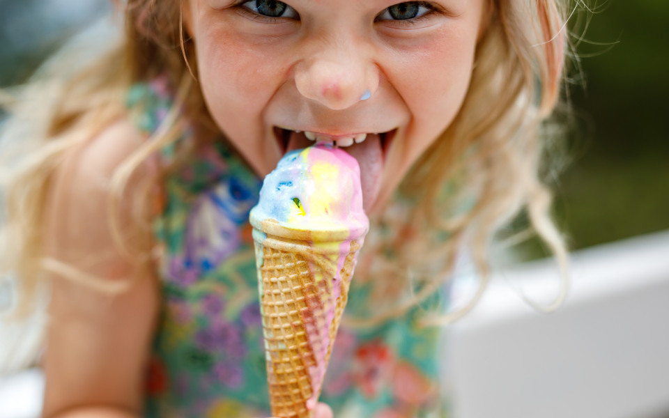 Ein kleines Mädchen mit blonden lockigen Haaren isst ein buntes Eis in einer Waffel.