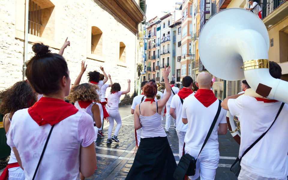 Menschen in weißer Kleidung mit roten Halstüchern tanzen und feiern auf einer engen Straße zwischen historischen Gebäuden, begleitet von einer Blaskapelle.