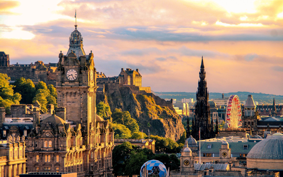 Panoramablick auf Edinburgh bei Sonnenuntergang mit dem markanten Uhrturm des Balmoral Hotels, dem Edinburgh Castle auf dem H&uuml;gel, dem gotischen Scott Monument und einem Riesenrad im Stadtzentrum.