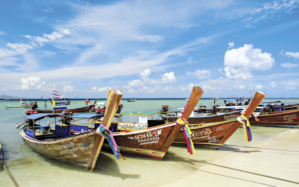 Traditionelle Longtail-Boote in Thailand am wei&szlig;en Sandstrand von Phuket, verziert mit bunten B&auml;ndern, vor t&uuml;rkisfarbenem Wasser und blauem Himmel mit Wolken.