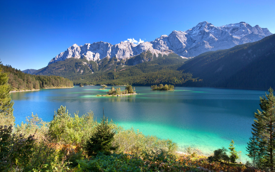 Blick auf den t&uuml;rkisfarbenen Eibsee mit kleinen Inseln, umgeben von dichtem Wald und den schneebedeckten Gipfeln der Zugspitze in Bayern, Deutschland.        ChatGPT fragen
