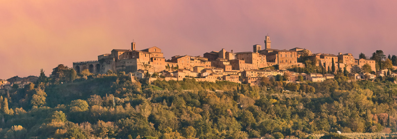Radfahrerin auf einem Feldweg in der Toskana, Italien, im warmen Abendlicht; im Hintergrund liegt die historische Stadt Pienza.